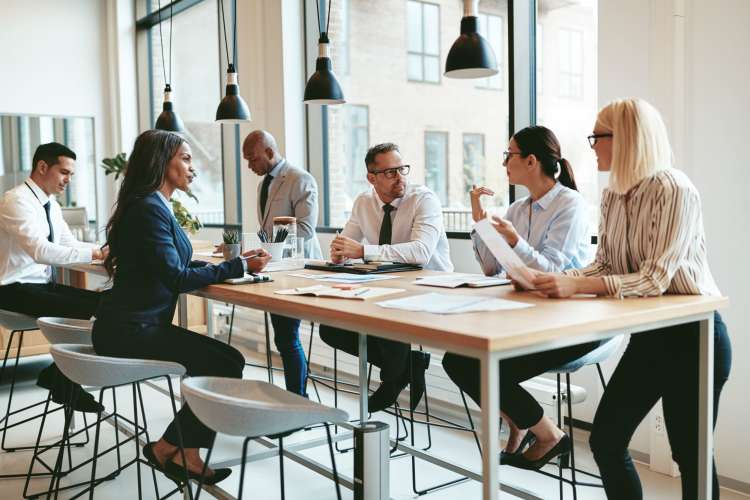 Six people having a discussion while seated and standing around a table at work