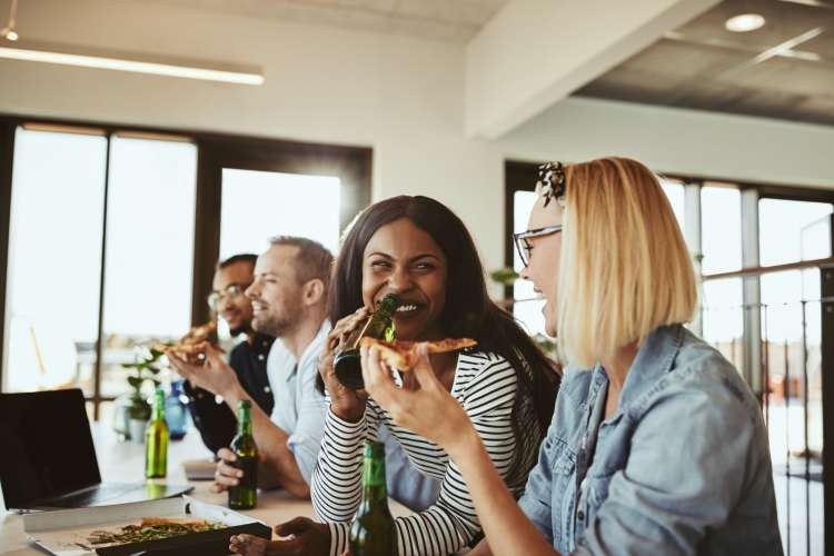 Diverse coworkers enjoying beer and pizza in their well-lit office lounge.