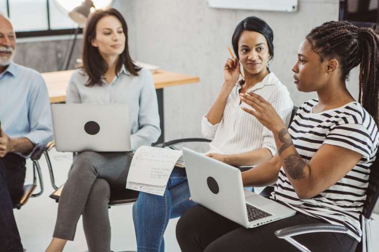 four coworkers sit in a circle with laptops and notes listening to a female colleague speak