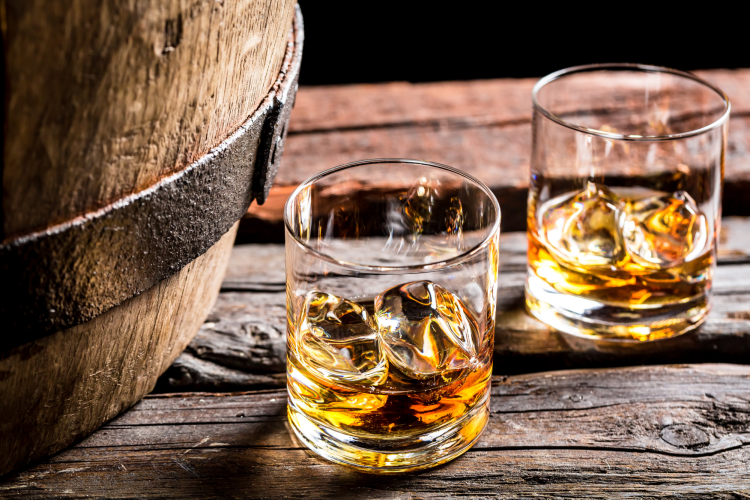 two glasses of whiskey on the rocks next to a bourbon barrel