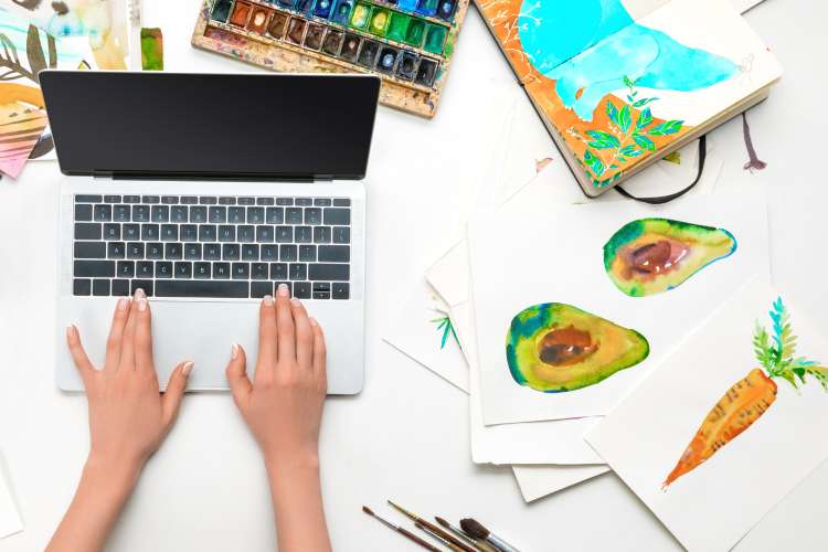 hands on laptop surrounded by watercolor paintings