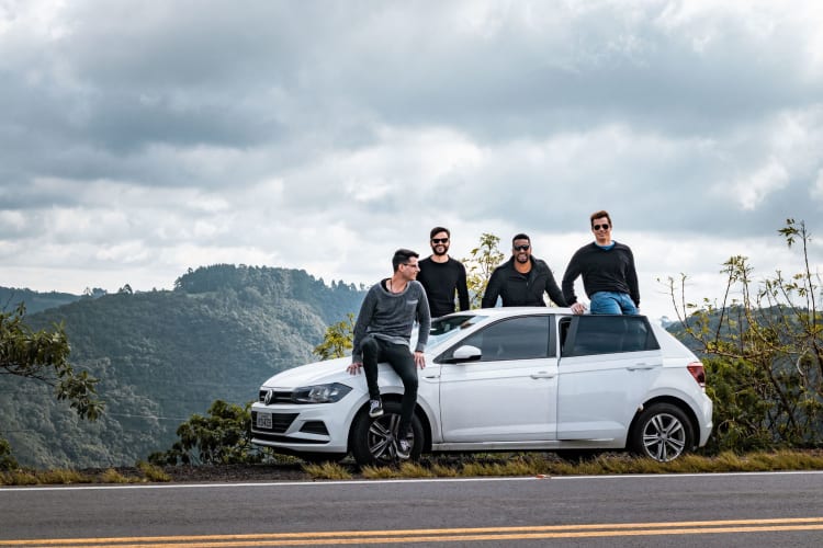 four men sit on or stand next to a car at the side of the road with a forest behind them