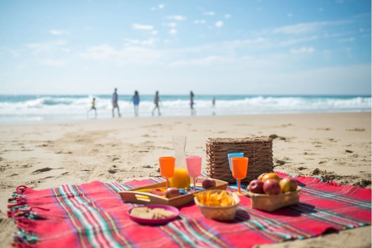 a red rug is laid out on the beach with picnic food while people play in the sea behind it