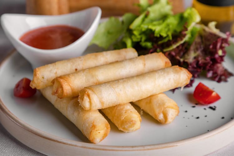 Six spring rolls on a plate with sauce and greens on the side