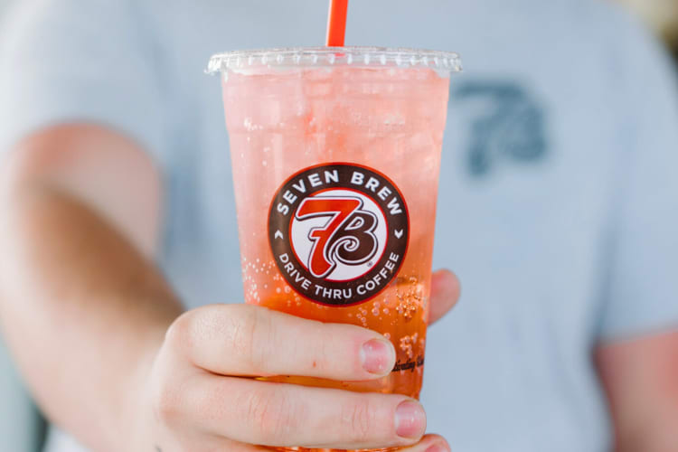 A person holding a peach colored drink in a clear take out cup