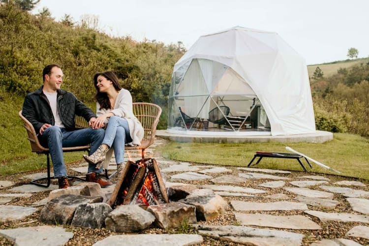 A couple sitting next to a campfire and a glamping tent