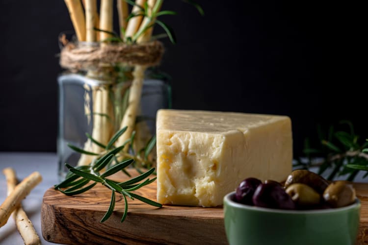 A block of Asiago on a board next to olives and bread sticks