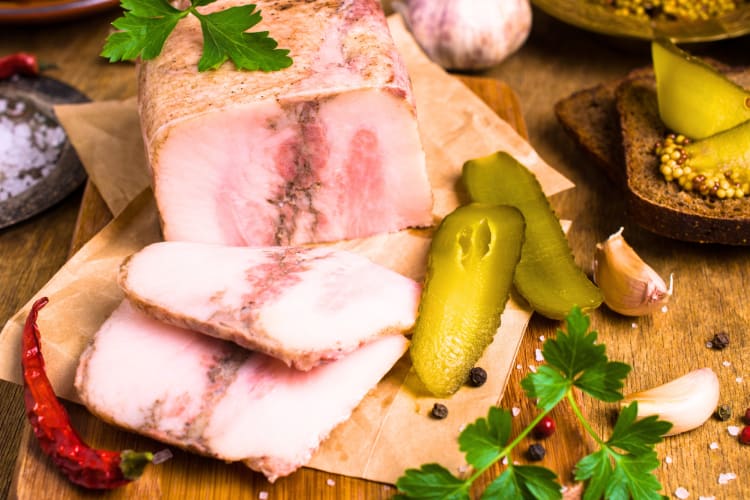 Lardo di Colonnata sliced on a chopping bored surrounded by pickles and other vegetables. 