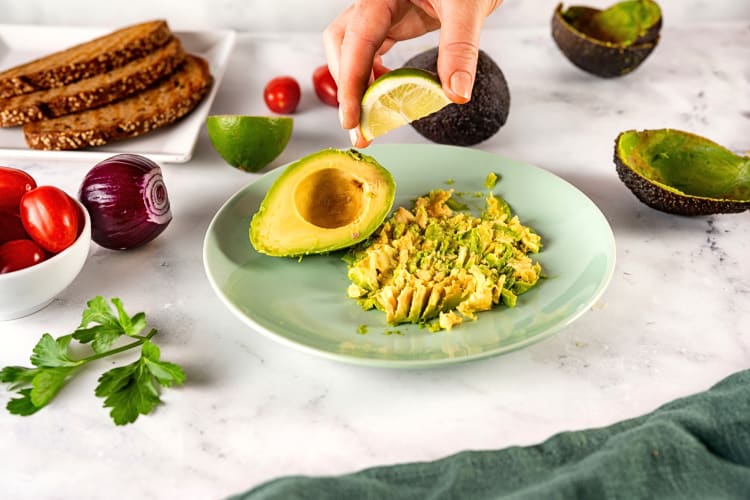 Hand squeezing a lemon wedge over mashed avocado, surrounded by bread, an onion and tomatoes