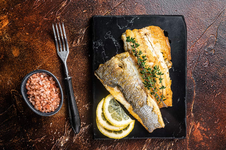 Aerial view of branzino and lemon slices on a black serving stone, next to a bowl of salt and a fork