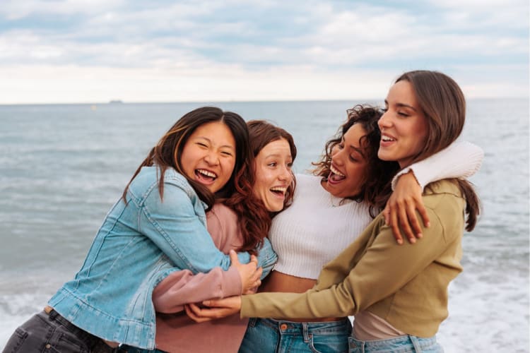 a group of women hug and laugh on the beach with the ocean behind them