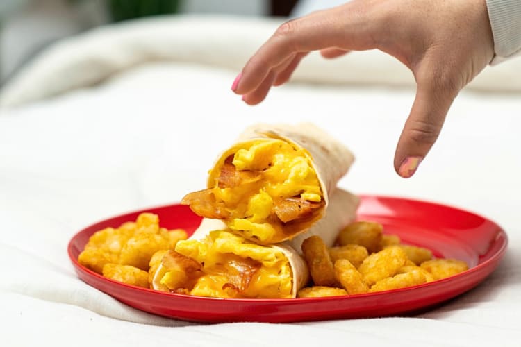 Person's hand reaching for a plate with two burritos and chicken pieces on it