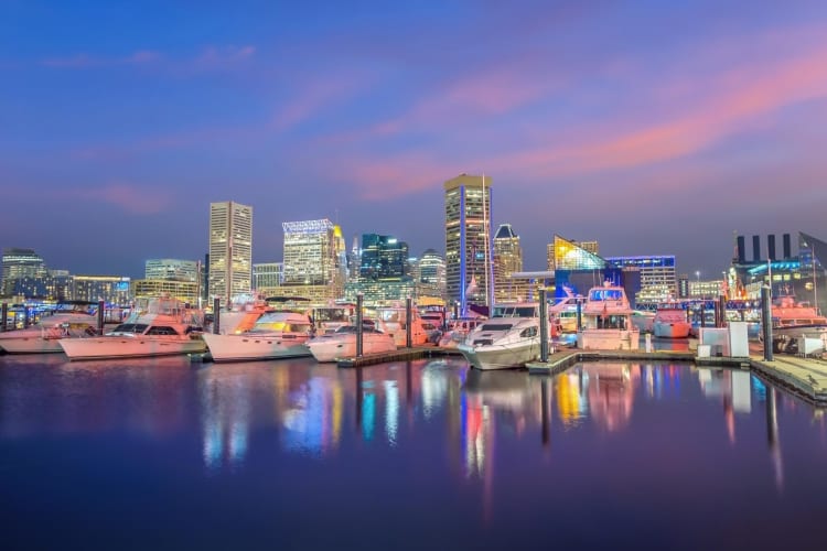 A harbor with boats on the water next to illuminated city buildings under a purple sky