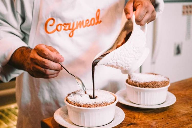 A person in an apron that says Cozymeal pouring chocolate sauce into souffles