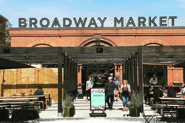 A brick building with a sign that says Broadway Market with tables and chairs in a courtyard in front