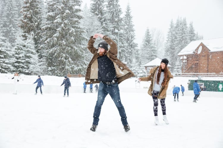 A couple ice skating near other people and trees as it snows