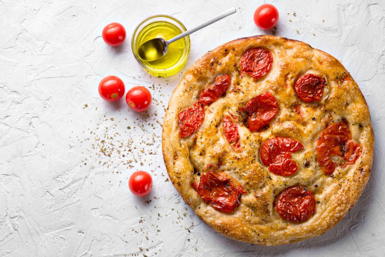A round focaccia with tomatoes next to fresh tomatoes and olive oil