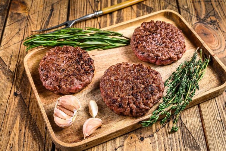 Three beef patties on a wooden chopping board with herbs and garlic
