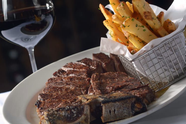 Sliced portion of steak with fries next to it on a plate, and a glass of wine in the background