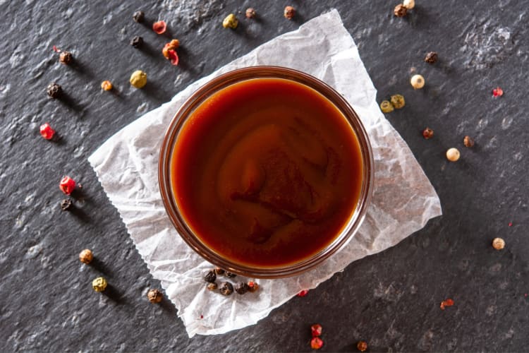 A small bowl of BBQ sauce surrounded by peppercorns