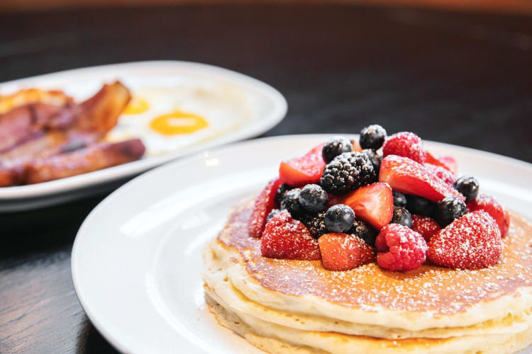 Pancakes with berries next to a plate with bacon on a table