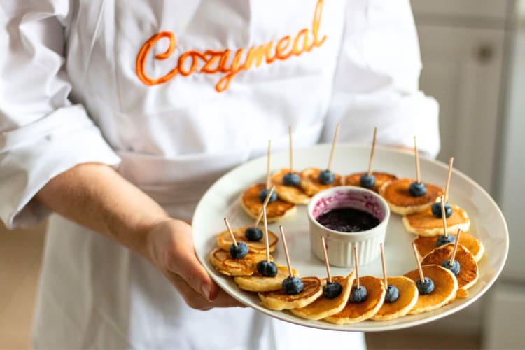 A person in a Cozymeal apron holding a plate of mini pancakes