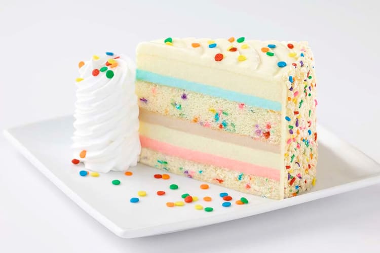 A colorful layered cake and cheesecake dessert with confetti sprinkles and whipped cream