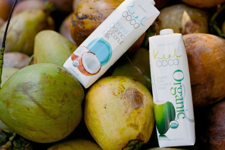 Two containers of coconut water next to fresh, young coconuts