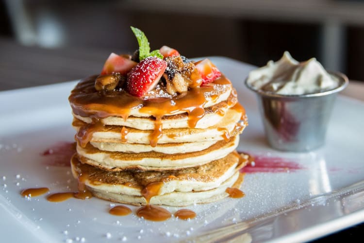 A beautifully presented stack of pancakes topped with syrup, diced fruit and a dusting of sugar.