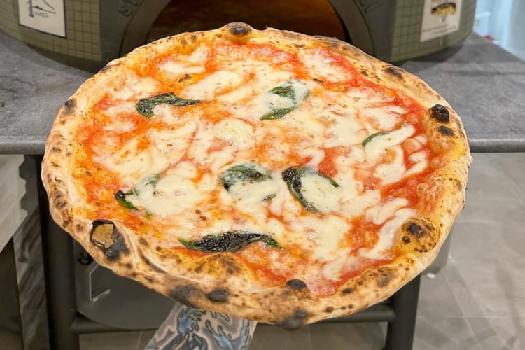 A person holding a pizza Margherita next to a pizza oven