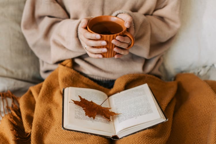 A person holding a cup of tea with a blanket and a book