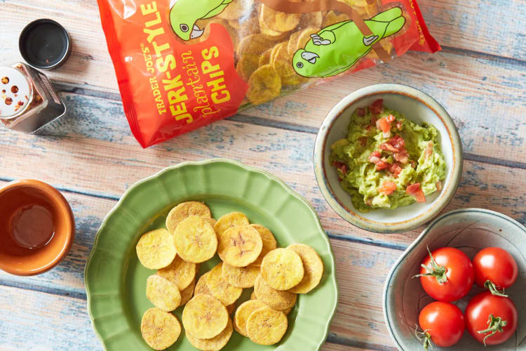 Plantain chips on a plate next to guacamole, tomatoes and a bag of plantain chips