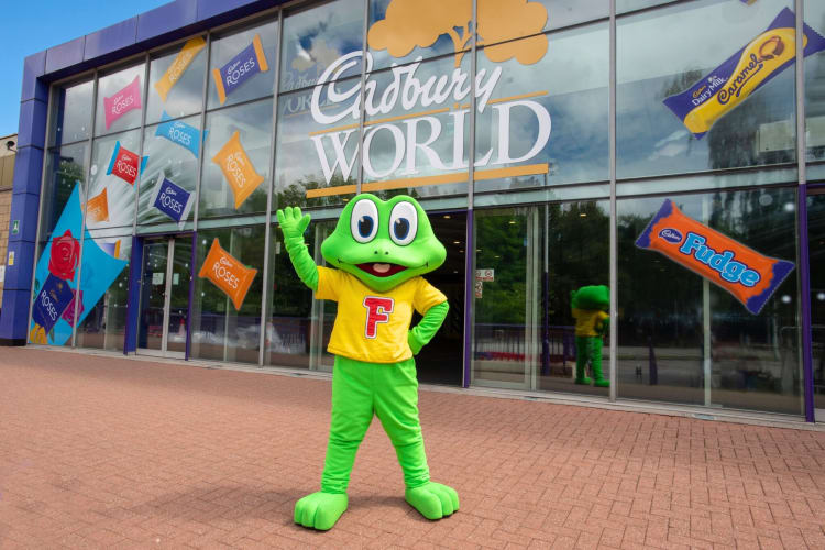 A frog mascot character in front of a glas Cadbury World building