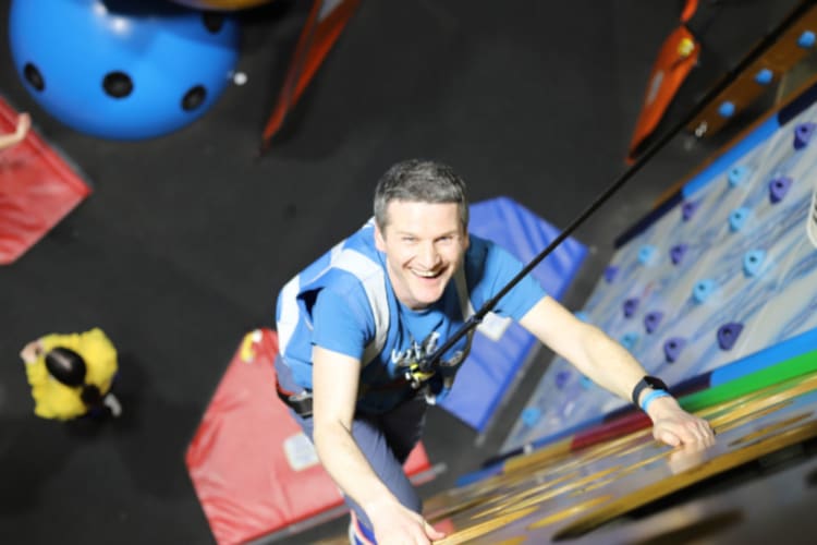 a man hangs from a rock climbing wall and smiles up at the camera