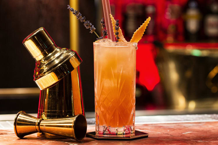 A gold cocktail shaker on a bar with an orange drink with lavender