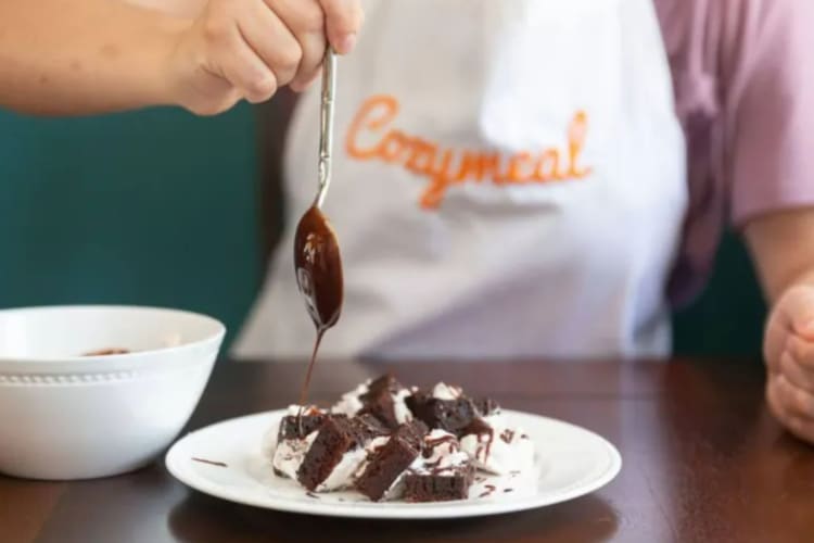 A person in a Cozymeal apron pouring chocolate sauce onto brownie bites