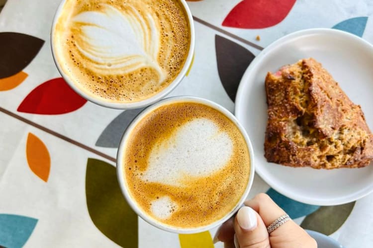 Two people holding cups of coffee with latte art next to a scone