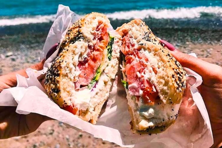 A person holding a filled bagel with the ocean in the background