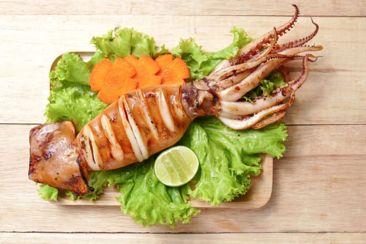 A whole squid cooked and presented on a wooden board with green salad, lime and carrots.