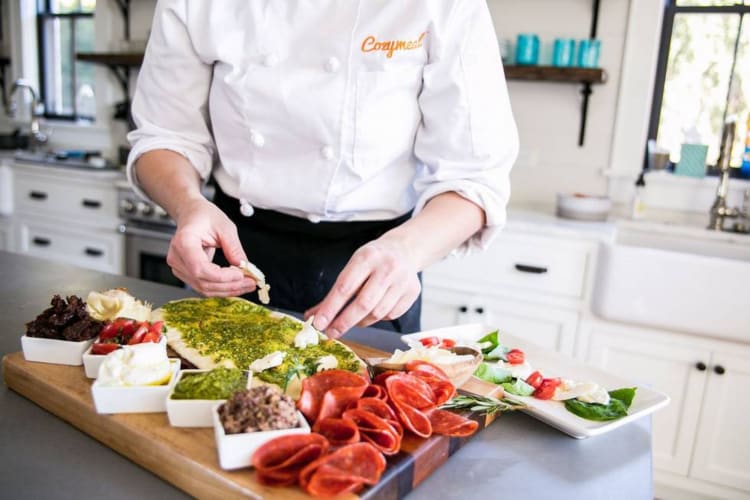 A chef in a Cozymeal apron preparing a charcuterie board