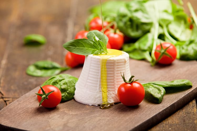 Ricotta cheese with oil being poured on it, surrounded by basil leaves and cherry tomatoes