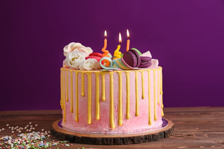 A birthday cake with pink and yellow frosting, macarons, icing flowers and lit candles.