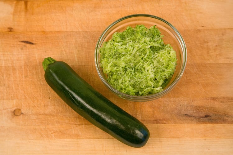 One whole zucchini next to a bowl of shredded zucchini