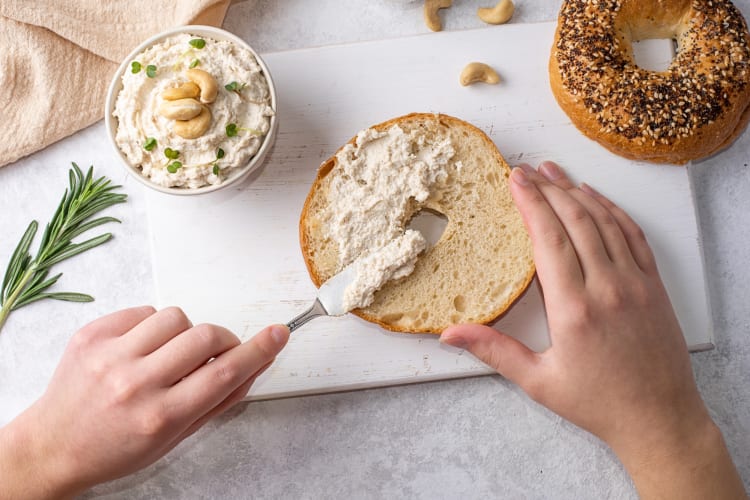 Aerial shot of a hand smearing cashew cream on a bagel, next to a bowl of cashew cream