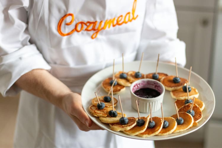 A chef in a Cozymeal apron holding a plate of mini pancakes