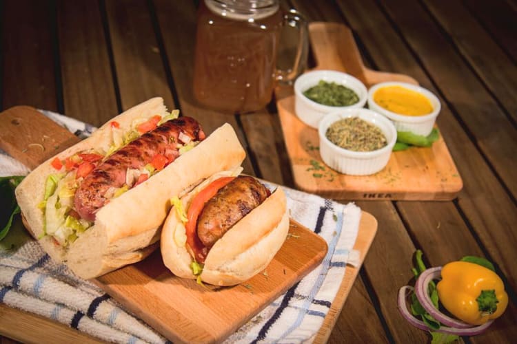 Two hot dogs on a chopping board in front of spices on another board from Che Grill