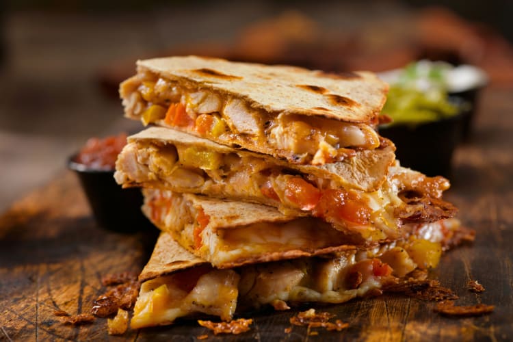 Cheese and chicken quesadilla on a wooden board with side sauces in the background