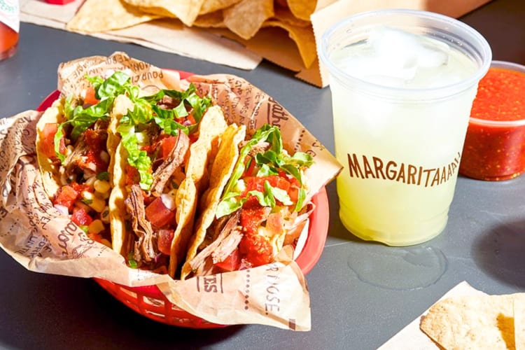 A basket of tacos next to a margarita in a take-out cup