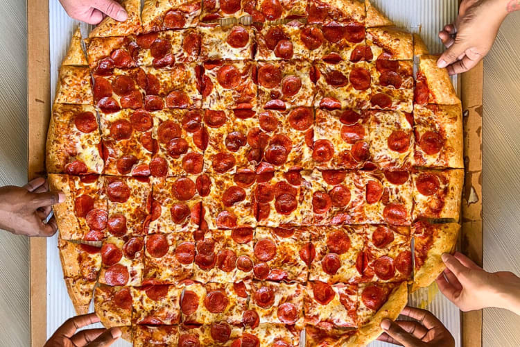 Aerial view of six hands grabbing square-cut slices from a large pizza