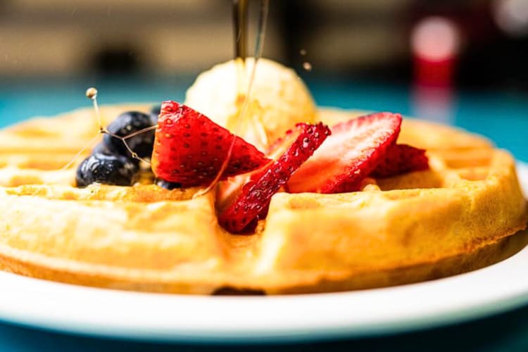 Eye-level view of syrup being poured over a waffle, topped with strawberries and blueberries